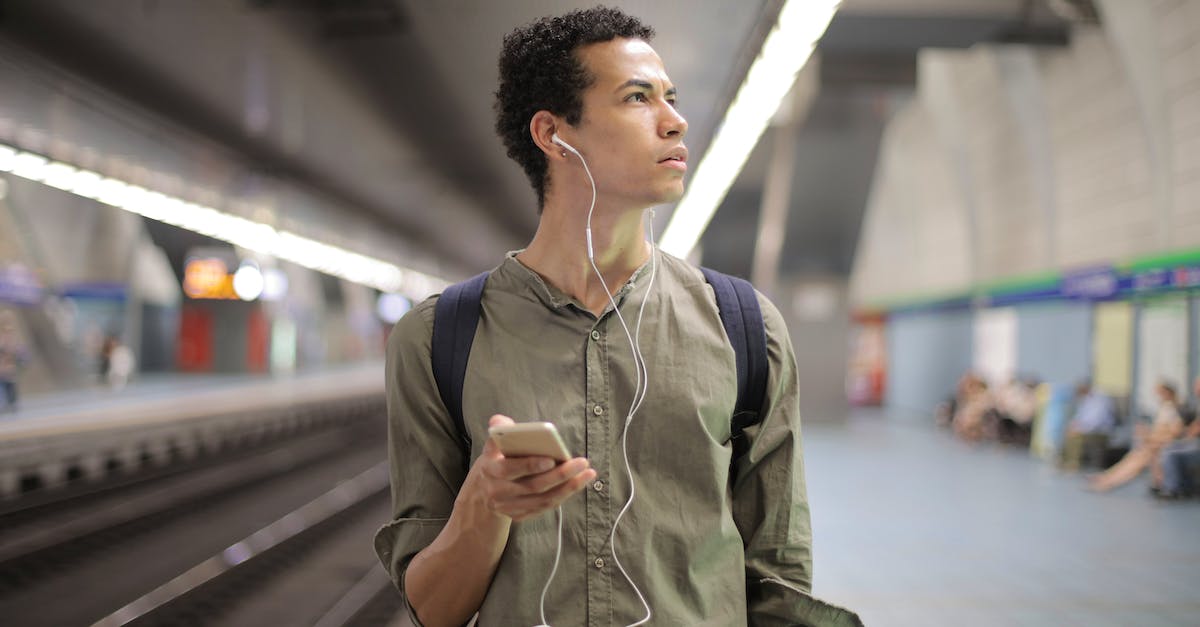 young ethnic man in earbuds listening to music while waiting for transport at contemporary subway st